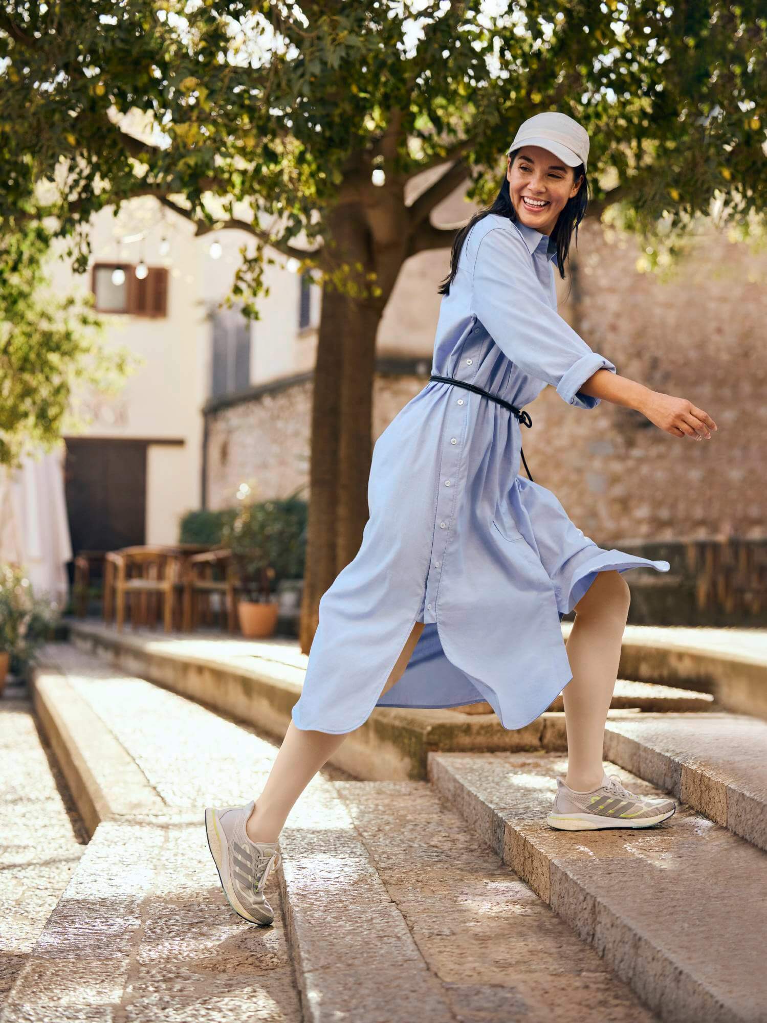 Eine lachende Frau mit dunklen Haaren trägt ein hellblaues Hemdkleid mit Gürtel, eine weisse Kappe und beige Turnschuhe. Sie bewegt sich dynamisch eine Steintreppe hinauf, während ihr Kleid im Wind flattert. Im Hintergrund sind ein Baum mit grünen Blättern, eine Steinmauer und ein Gebäude mit Fenstern und Holzbalken zu sehen. Die Szene wirkt lichtdurchflutet und idyllisch.